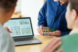 Ingénieur présentant une maquette de maison et des plans à des clients lors d’une étude technique avant réhabilitation.